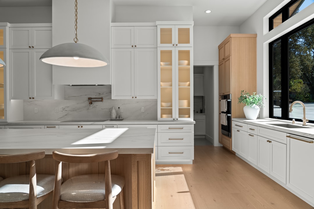 Bright modern kitchen with white shaker cabinets, warm wood accents, and marble backsplash. Contemporary custom home featuring a large island, brass hardware, statement pendant lighting, and black-framed windows. Luxury residential interior blending timeless design with natural materials and abundant light.